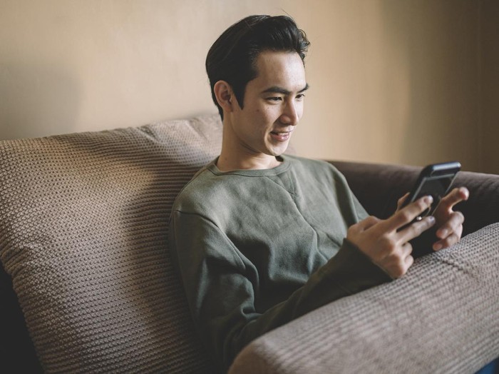an asian chinese man sitting on his sofa with cushion relaxing with his smart phone surfing the net and shopping online