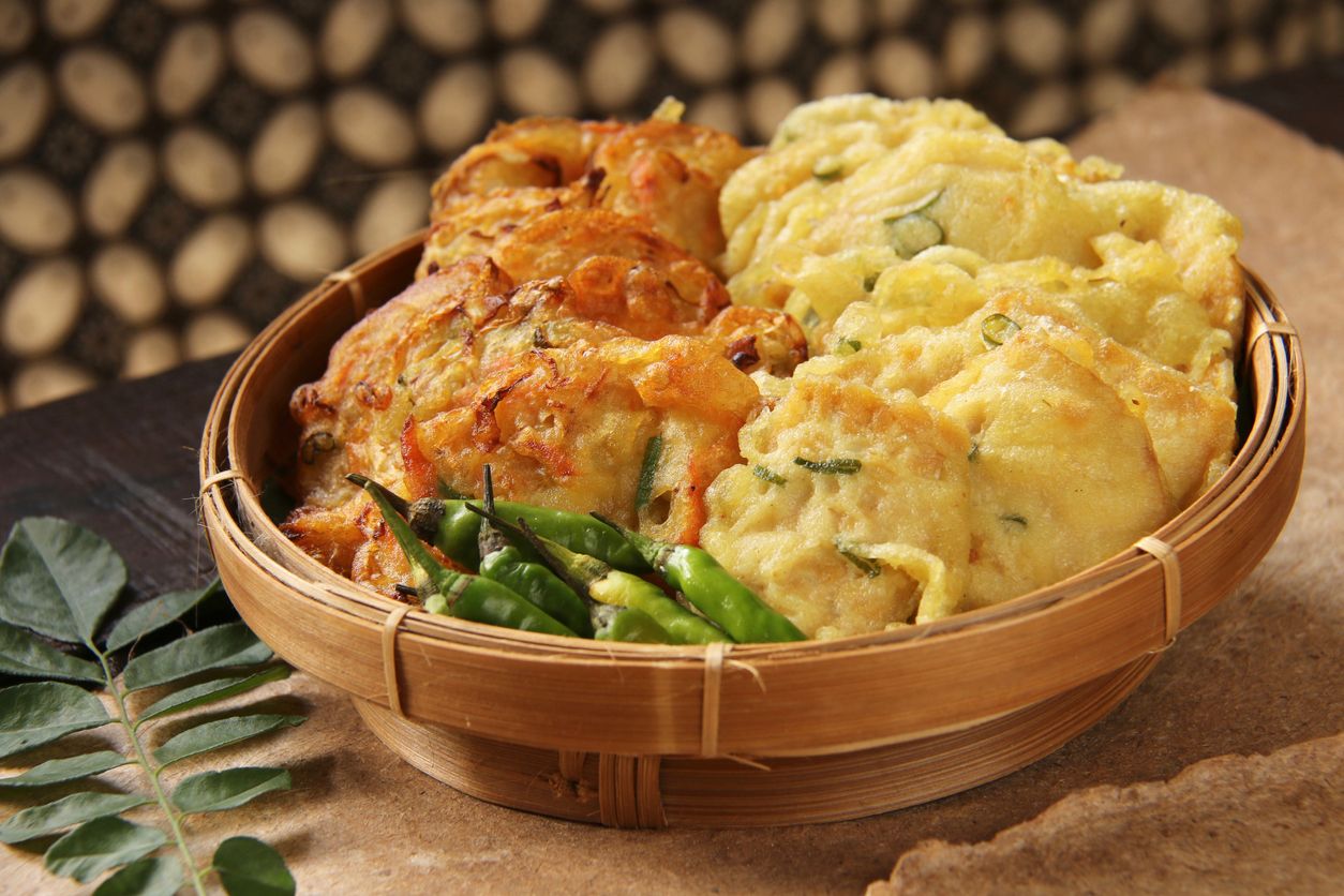 Gorengan, the popular and traditional Javanese side dish of food deep-fried in batter; a typical dish served in Nasi Kucing fare. This fritter dish consists of mixed vegetable fritters on the left and tempeh fritters on the right. Fritters are served with small green chili peppers. The dish is served on a woven bamboo plate lined with banana leaf. It is placed on a wooden table lined with recycled brown paper. In the background is a Javanese batik curtain.