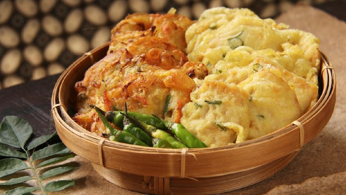 Gorengan, the popular and traditional Javanese side dish of food deep-fried in batter; a typical dish served in Nasi Kucing fare. This fritter dish consists of mixed vegetable fritters on the left and tempeh fritters on the right. Fritters are served with small green chili peppers. The dish is served on a woven bamboo plate lined with banana leaf. It is placed on a wooden table lined with recycled brown paper. In the background is a Javanese batik curtain.