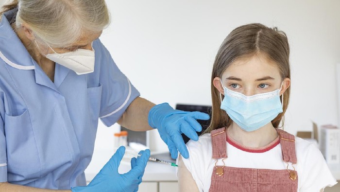 Ilustrasi anak mendapatkan vaksin A medium shot of a Caucasian, young girl receiving her Covid-19 vaccination from a nurse. They are both wearing a protective face mask.