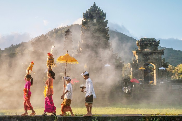 A mother and a teenaged girl are dressed in brightly colored sarongs, blouses, and sashes and are balancing tall fruit baskets on their heads. Two sons are dressed in sarongs and white shirts. The family is walking in front of an old stone temple building which has a smoky atmosphere.