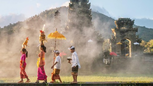 A mother and a teenaged girl are dressed in brightly colored sarongs, blouses, and sashes and are balancing tall fruit baskets on their heads. Two sons are dressed in sarongs and white shirts. The family is walking in front of an old stone temple building which has a smoky atmosphere.