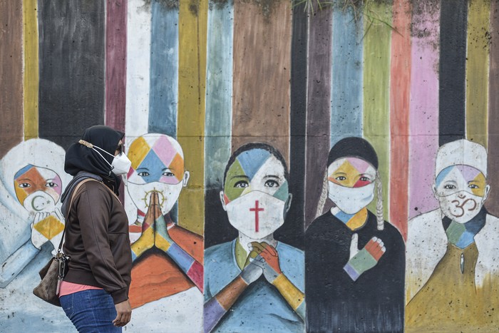 Seorang warga dengan mengenakan masker berjalan di depan mural imbauan mengenakan masker dari lintas agama di kawasan Jakarta Timur.