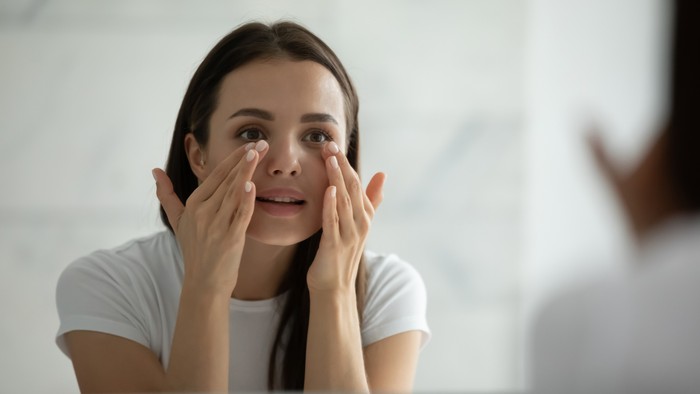Ilustrasi Krim Mata Close up smiling young woman wearing white t-shirt doing facial massage, applying moisturizing cream on under eye skin, looking in mirror, standing in bathroom, enjoying skincare procedure