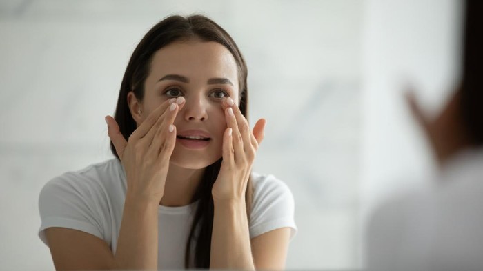 Ilustrasi Krim Mata Close up smiling young woman wearing white t-shirt doing facial massage, applying moisturizing cream on under eye skin, looking in mirror, standing in bathroom, enjoying skincare procedure