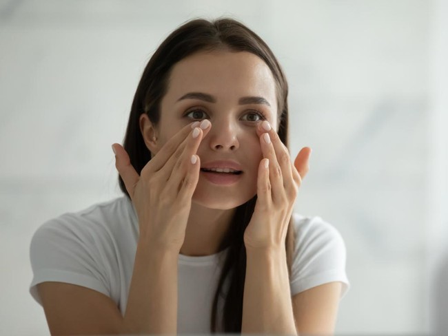Close up smiling young woman wearing white t-shirt doing facial massage, applying moisturizing cream on under eye skin, looking in mirror, standing in bathroom, enjoying skincare procedure