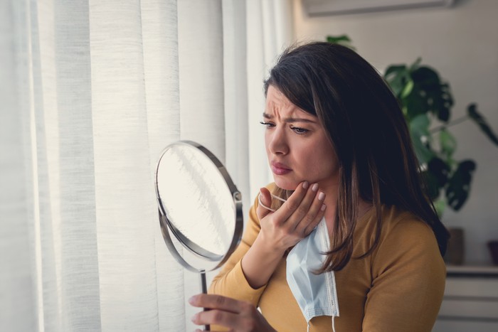 Ilustrasi Bekas Jerawat Woman looking herself in the mirror at home standing by the big window. She is concerned about acne, maskne