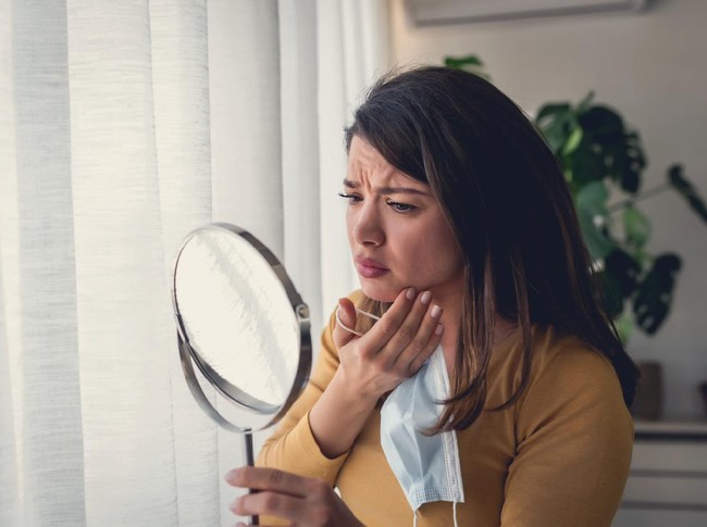 Ilustrasi Bekas Jerawat Woman looking herself in the mirror at home standing by the big window. She is concerned about acne, maskne