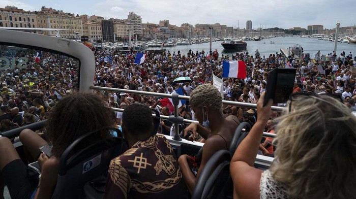 Warga Prancis Kembali Protes Aturan Kartun Vaksin Tourists watch a protest from a sightseeing bus in Marseille, southern France, Saturday, Aug. 7, 2021. Thousands of people are marching in Paris and other French cities during a fourth consecutive week of protests against COVID-19 entrance requirements. The demonstrations on Saturday come days after France's Constitutional Council upheld most provisions of a new law that expands the locations where health passes are needed. (AP Photo/Daniel Cole)