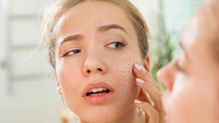 Young beautiful woman touching skin in bathroom. Unhappy girl standing in towel, looking in the mirror, checking dry irritated skin. Morning skincare routine.
