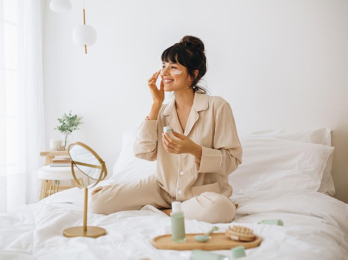 Ilustrasi Skincare Happy woman doing routine skin care at home with beauty products. Woman sitting on bed at home and applying face cream.