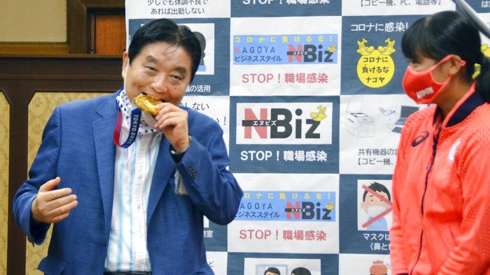 Nagoya Mayor Takashi Kawamura, left, bites the Olympic gold medal of Miu Goto, right, of Japan softball team who won the event at the 2020 Summer Olympics, at the city office building in Nagoya, central Japan, Wednesday, Aug. 4, 2021. (Kyodo News via AP)
