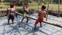 Memanfaatkan sepetak kolam berdasar lumpur, dua orang bocah duduk di atas sebatang bambu sambil mengayunkan bantal guling pada teman di hadapannya.