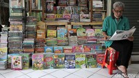 Seorang pedagang menunggu pembeli dengan membaca koran di depan kiosnya.