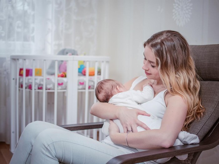 Breastfeeding baby near crip. Pretty mother holding her newborn child. Mom nursing baby. Beautiful woman and new born relax at home in nursery interior. Blond mother breast feeding baby.