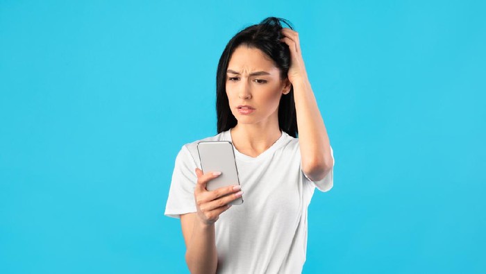 Dissatisfied gloomy female model looking at smartphone while browsing social media or texting sms and being disappointed, standing over blue background. Woman trying to call but there is no signal