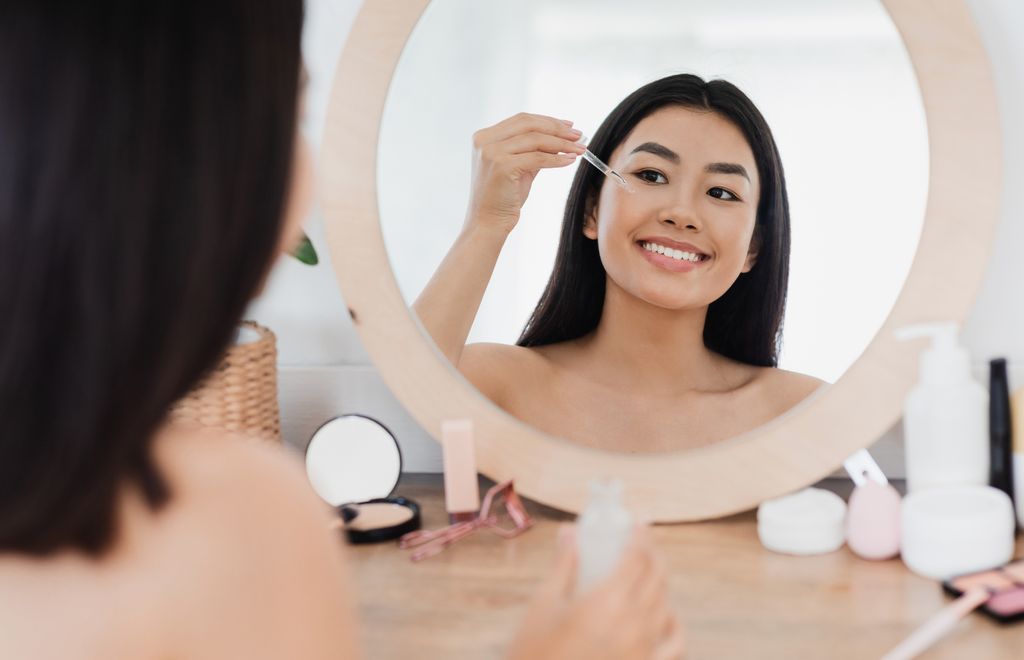 Eye zone care concept. Happy beautiful asian girl applying eye serum in front of mirror at home