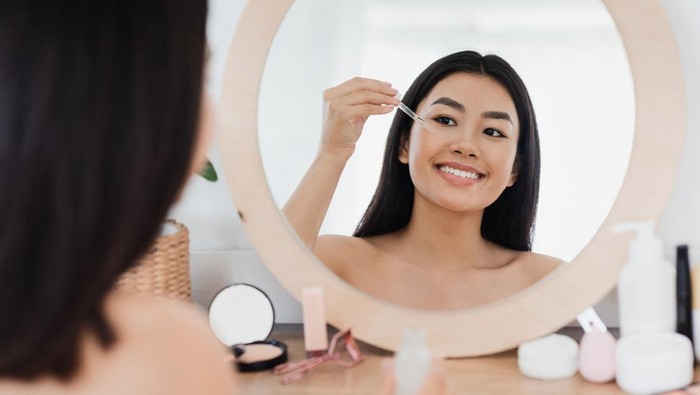 1265941373 Eye zone care concept. Happy beautiful asian girl applying eye serum in front of mirror at home