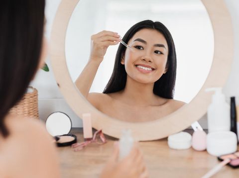 Eye zone care concept. Happy beautiful asian girl applying eye serum in front of mirror at home