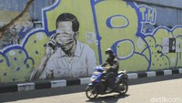Dari pantauan detikcom, Rabu (25/8/2021) sesosok pria yang tergambar dalam mural tersebut berambut poni dan menggunakan baju kemeja berwarna putih. Terlihat sosok pria tersebut tengah berpose sambil memegang kening sebelah kanan.  