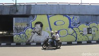 Pengendara motop melintas di depan mural mirip Jokowi dengan mata tertutup masker di Jalan Prabu Dimuntur, Kota Bandung, Kamis (26/8/2021). Terlihat sejumlah petugas masih mengerik kertas sisa gambar tersebut, kemudian memberikan cat putih.  