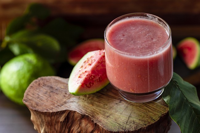 Delicious red guava juice alongside a slice of guava, in the background guavas and leaves on rustic wood