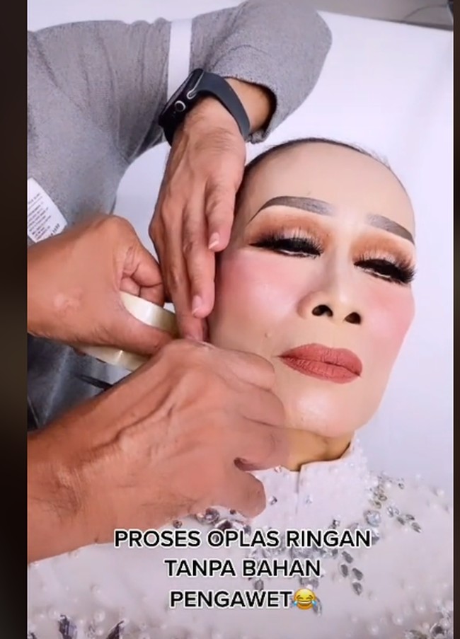 Kemudian masuk ke proses yang bikin netizen terheran-heran. Rangga memperlihatkan cara dia memasang lakban di wajah sang nenek. Bernada guyon, Rangga menyebut proses tersebut sebagai oplas ringan tanpa bahan pengawet. Dari rahang, lakban ditarik di balik kuping. Proses penarikan wajah dengan lakban ini sukses membuat wajah sang nenek jadi lebih kencang. Hasilnya, wajah nenek tampak manglingi. Foto: dok. TikTok/@ranggajuans