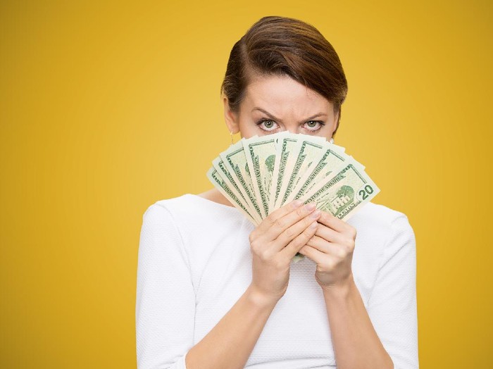 1064696264 Young grumpy woman covering face with heap of bills looking at camera on yellow background