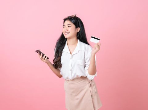 Young smiling Asian woman holding credit card with smartphone and looking to copy space on pink background.