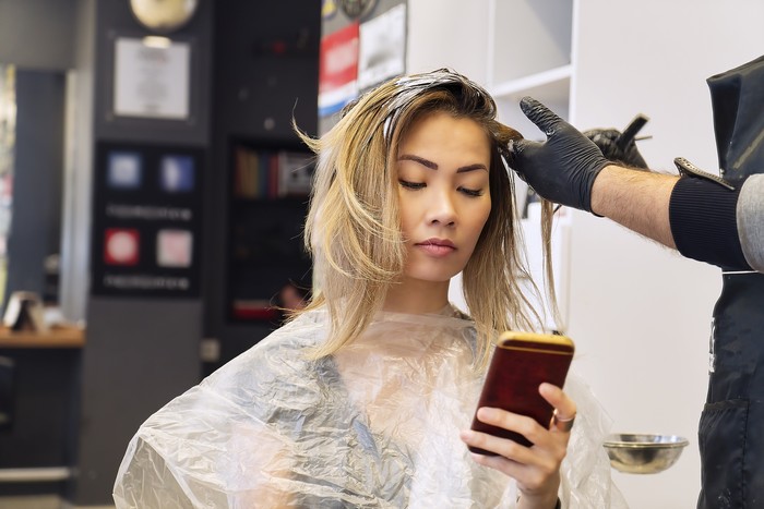 1088633502 Woman on mobile phone when having hair coloring process in a hair salon