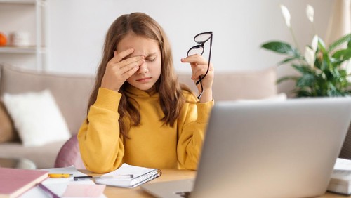 Tired school girl rubbing her eyes, holding eyeglasses, exhausted from studying at home and doing homework, sitting in front of laptop with notebook. Homeschooling, distance education due to covid-19