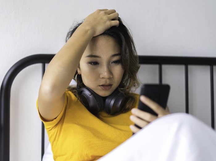 Foto ilustrasi wanita yang mengalami ghosting. Stressed Asian teenager receiving bad news on mobile phone, on her bed.