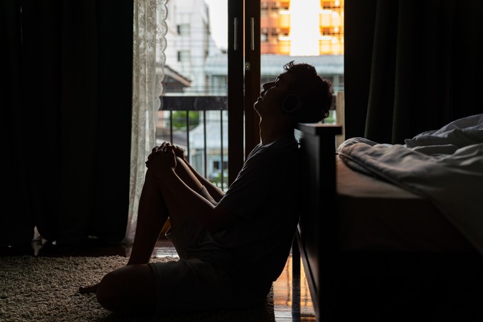 Wide shot young adult asian loneliness sad man sitting on the floor in bedroom leaning on bed and cry. Broken heart despair man sitting on the floor listening music from headphone alone.