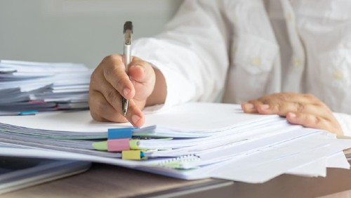 Teacher hand is holding pen for checking student homework assignments on desk in school. Unfinished paperwork stacked in archive with color paper and binder paper clips. Education and business concept