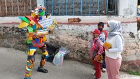 Pria berkostum robot itu mendekati siswa SD yang kebetulan sedang ditunggu oleh orangtuanya. Ternyata ia membagikan sepaket buku dan alat tulis bagi siswa tersebut diselipi dengan sosialisasi protokol kesehatan COVID-19. 
