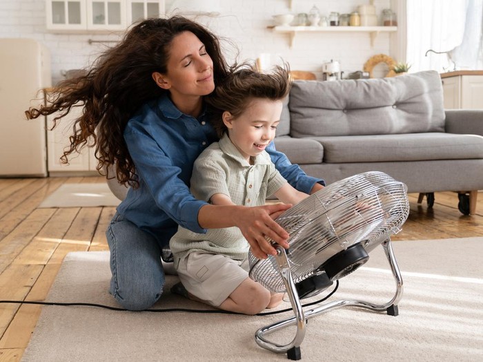 Cheerful young mom or nanny have fun with preschool boy at home sitting together in front of big fan dlowing cooling wind in living room. Carefree caring woman of 30 spend time with little son bonding
