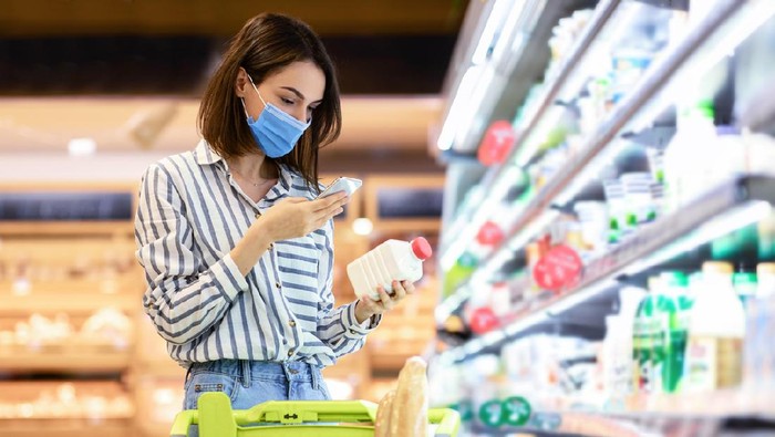 Ilustrasi Pasar Young woman in disposable face mask taking dairy products from shelf in the supermarket, holding bottle and smartphone, scanning bar code on product through mobile phone, walking with trolley cart
