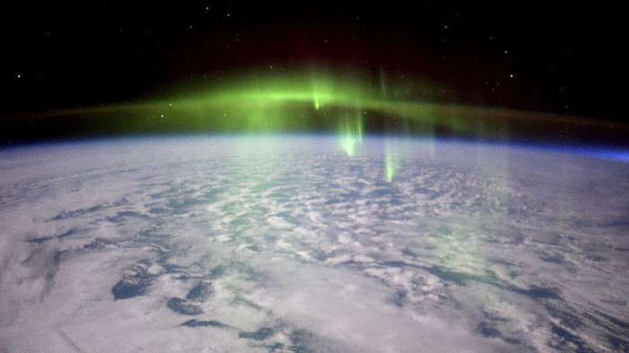 Penampakan spektakuler aurora seakan menyelimuti Bumi, dari titik 240 kilometer di atas sana memperlihatkan cahaya hijau aurora tinggi di atmosfer sementara di kejauhan, ada sinar kemerahan. Sedangkan di bawahnya adalah area awan-awan yang berarak di atas lautan.