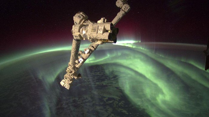 Penampakan spektakuler aurora seakan menyelimuti Bumi, dari titik 240 kilometer di atas sana memperlihatkan cahaya hijau aurora tinggi di atmosfer sementara di kejauhan, ada sinar kemerahan. Sedangkan di bawahnya adalah area awan-awan yang berarak di atas lautan.