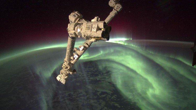 Penampakan spektakuler aurora seakan menyelimuti Bumi, dari titik 240 kilometer di atas sana memperlihatkan cahaya hijau aurora tinggi di atmosfer sementara di kejauhan, ada sinar kemerahan. Sedangkan di bawahnya adalah area awan-awan yang berarak di atas lautan.