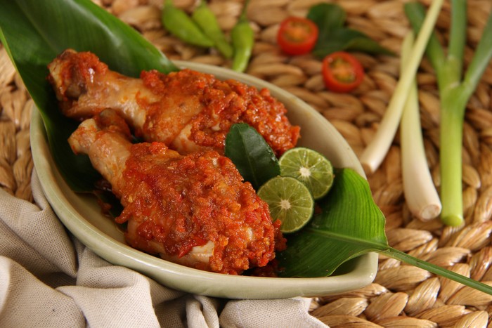 Ayam Rica-Rica, the popular and traditional dish from Manado, chicken braised in spicy red chili paste. Two drumsticks are arranged on a small ceramic plate that has been lined with a banana leaf. Two lime halves and a lime leaf are used to garnish the dish. The plate is placed on a woven bamboo place mat. Some of the herbs and spices are also placed on the place mat.