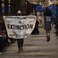 Di tengah acara, tiba-tiba muncul seorang perempuan membawa spanduk bertulisan Overconsumpstion = Extinction. Seakan menjadi bagian dari acara tersebut, ia berjalan mengikuti alur jalan para model. (Foto: Vianney Le Caer/Invision/AP)