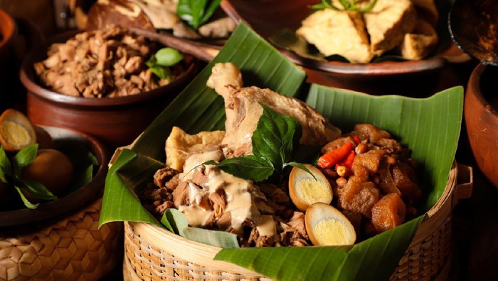 Gudeg Jogja, a Javanesse signature dish of young jackfruit stew; accompanied with white chicken curry and spicy stew of cattle skin cracker. All the dishes are arranged in a bamboo basket as a take-out container. In the background is each of the individual dish that makes up Gudeg Jogja.