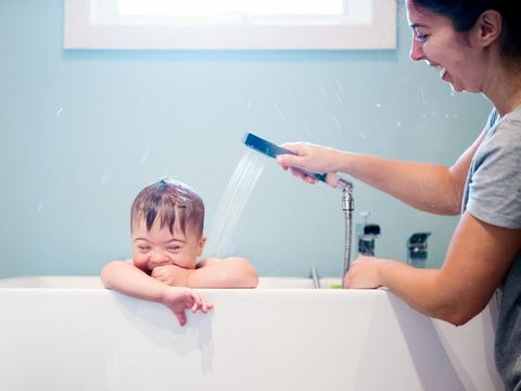 Down's syndrome sweet boy in the bath while Mom is washing him. He has blue eyes and blond hair. He is sitting in the bath and looking directly the camera. Mom watering her son with the shower. He as a cute face. The color and horizontal Photo was taken in Quebec Canada. There is copy space in this picture.