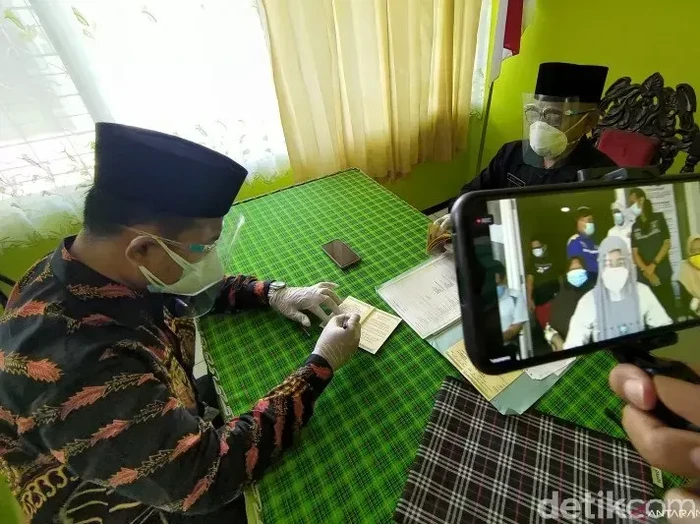 Mempelai pria menandatangani buku nikah disaksikan penghulu dan mempelai perempuan yang mengikuti prosesi akad nikah secara daring di Kantor Urusan Agama Kecamatan Pakel, Tulungagung, Jawa Timur, Sabtu (26/12/2020). Mempelai pria menandatangani buku nikah disaksikan penghulu dan mempelai perempuan yang mengikuti prosesi akad nikah secara daring di Kantor Urusan Agama Kecamatan Pakel, Tulungagung, Jawa Timur, Sabtu (26/12/2020).