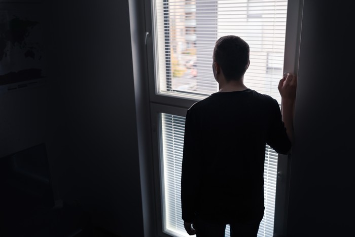 Lonely man looking out the window. Solitude, isolation and loneliness concept. Sad upset person with regret, shame or stress in moody dramatic back view silhouette. Dark room with shadow at home.