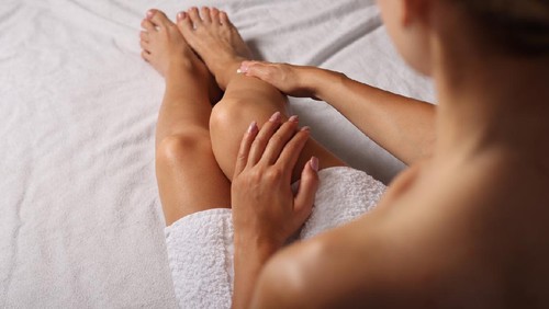 Attractive woman applying healthy cream on legs and sitting in bed after bathing. Girl wears white towel. Skincare and beauty concept