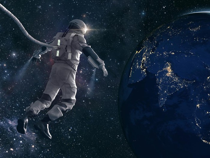 An CG astronaut in a modern space suit, connected to a tethered lifeline floats in deep space and looks at the lights of planet earth as the sun rises. Distant stars and galaxies are visible in the background. Credit: NASA https://earthobservatory.nasa.gov/images/79790/city-lights-of-asia-and-australiaand ESO for background images.