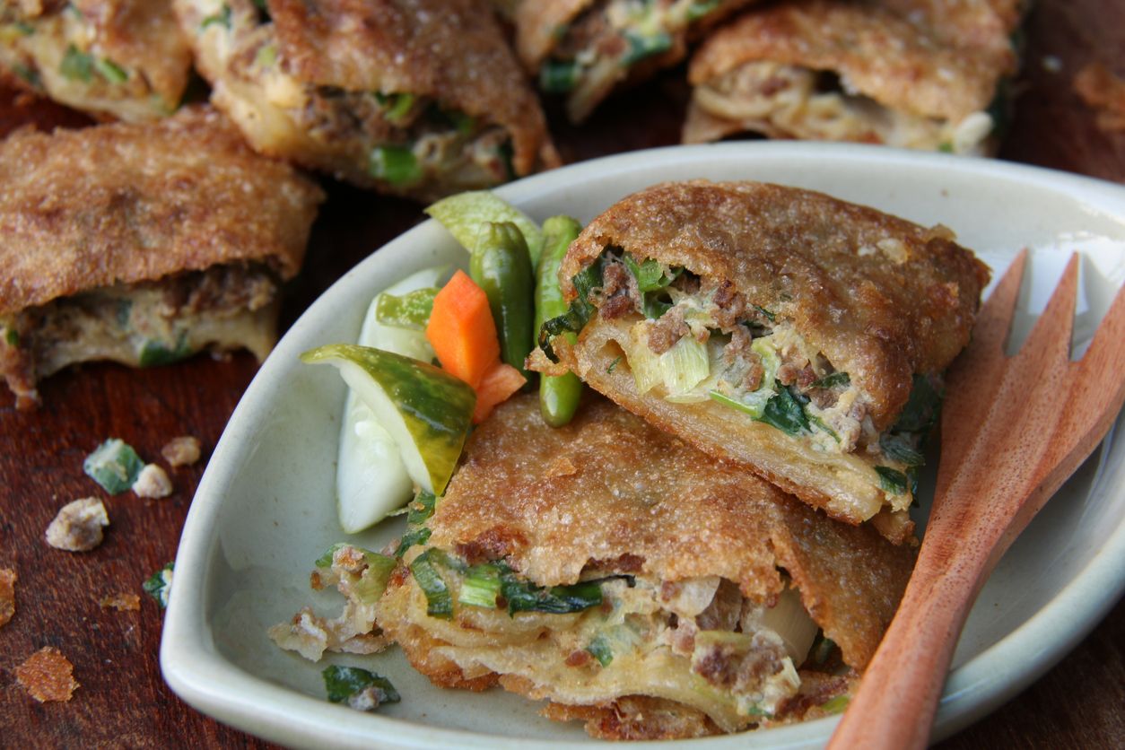 Martabak Telor, or also known as Martabak Malabar, is a pan-fried pastry cake stuffed with egg, ground meat, green onion, and spices. Here the martabak has been cooked and cut into squares; two pieces are plated and accompanied with spicy cucumber pickle. A wooden fork is placed on the right hand side of the served martabak pieces. Further back in the background, some martabak pieces are shown in blur.