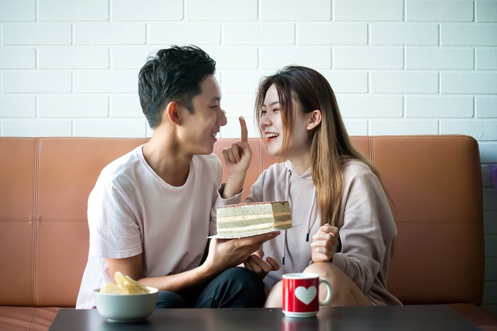 Beautiful Girl Surprise Her Boyfriend With Gift Celebrating Anniversary While He Is Sitting on the Sofa in the Living Room. Romantic Surprise for Husband Boyfriend, Happy Birthday Concept
